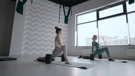 Group-of-young-women-stretching-in-gym-with-windows.-Two-attractive-sport-girls-work-out-nauka-asana-boat-yoga-pose-on-black-mat-in-fitness-class.-Two-women-stretching-legs-together-in-yoga-studio